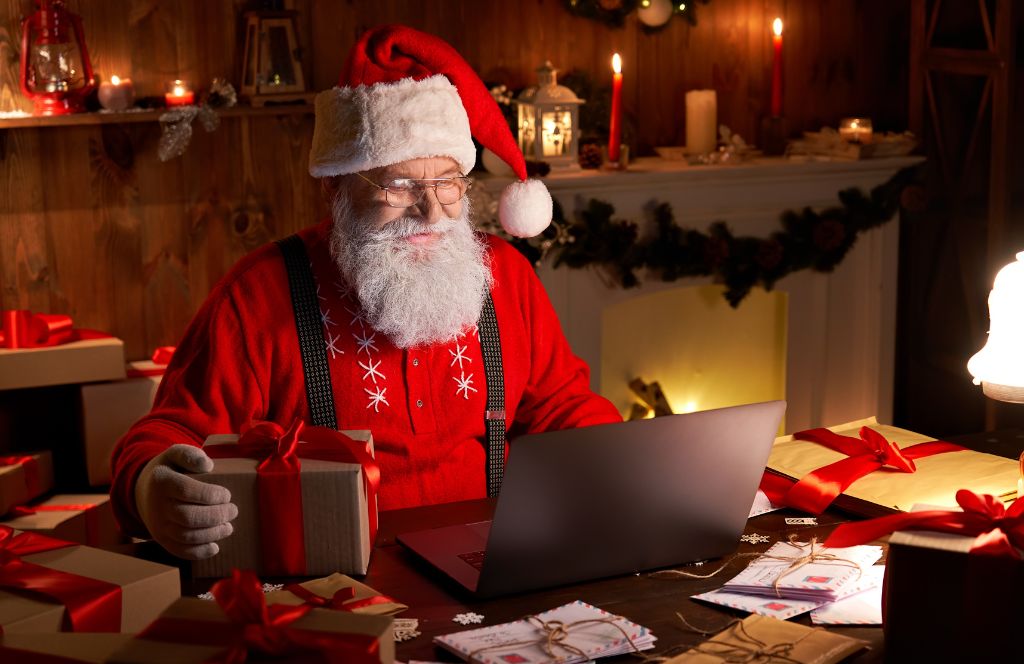 een man met een kerstmuts en bril die aan een tafel met een laptop zit